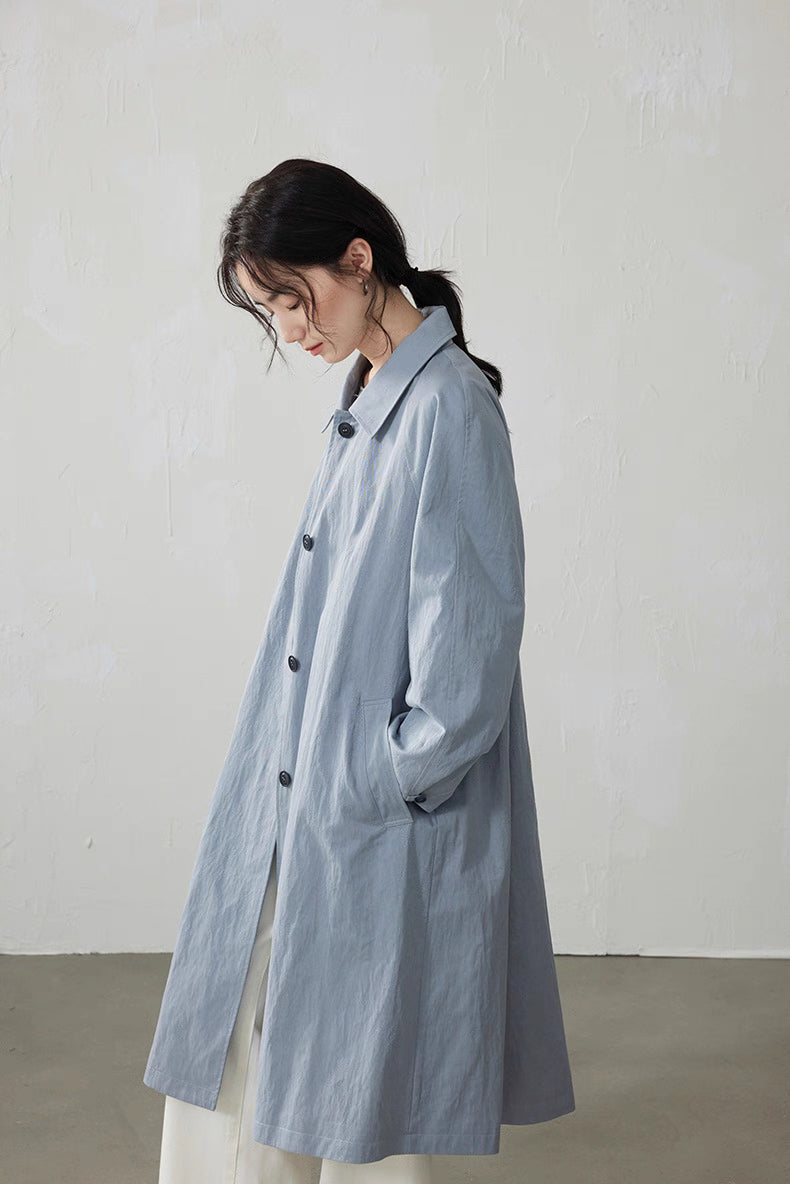 Side view of light blue-gray A-line trench coat showing raglan sleeves and relaxed silhouette.