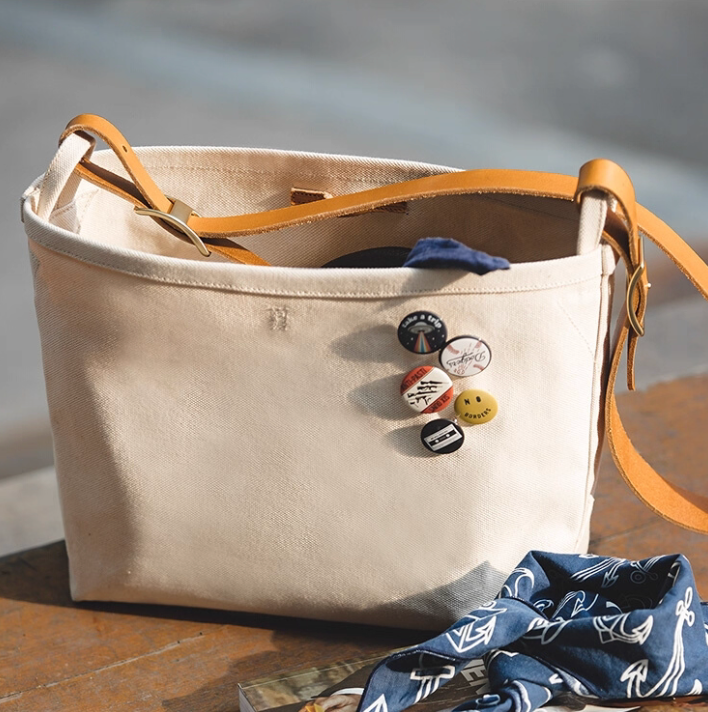 Natural canvas tote with leather strap decorated with enamel pins on table