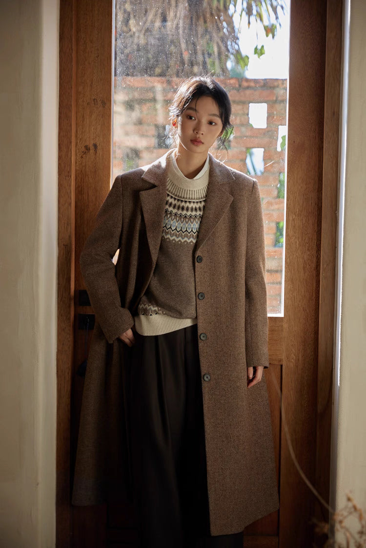Model in a brown herringbone long coat styled with a cream patterned sweater near a sunlit doorway.