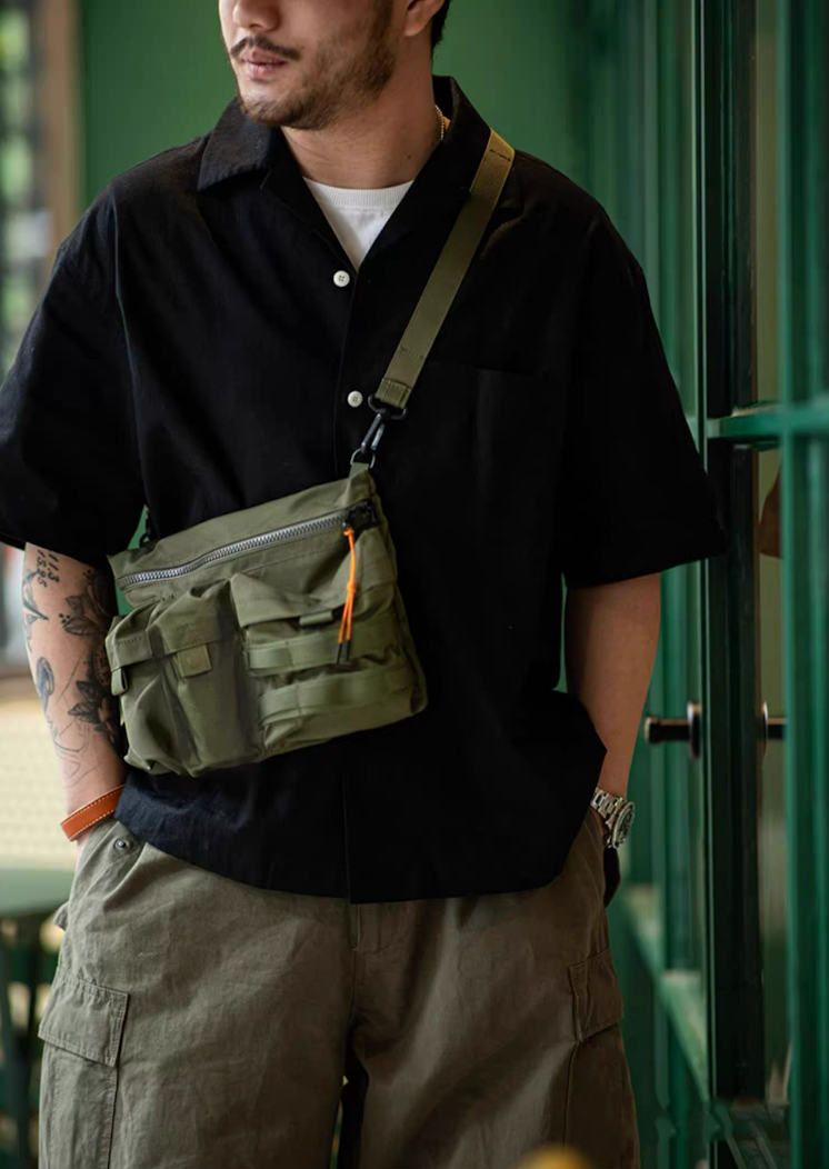 Man wearing army green crossbody bag with black shirt and khaki pants in urban setting