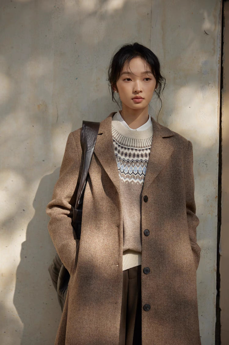 Woman in a brown herringbone wool coat standing outdoors in gentle sunlight against a warm wall.