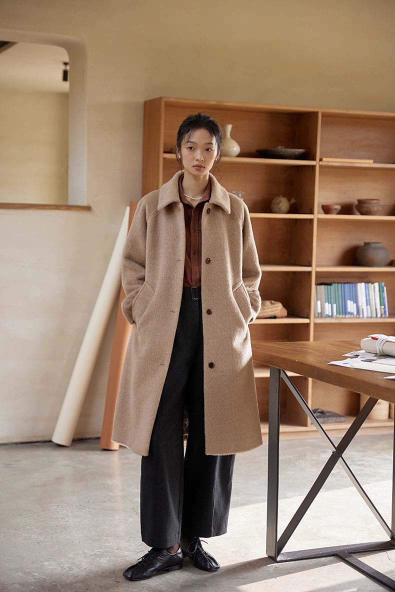 Full-body view of woman wearing oat beige Suri alpaca long coat in an art studio setting.