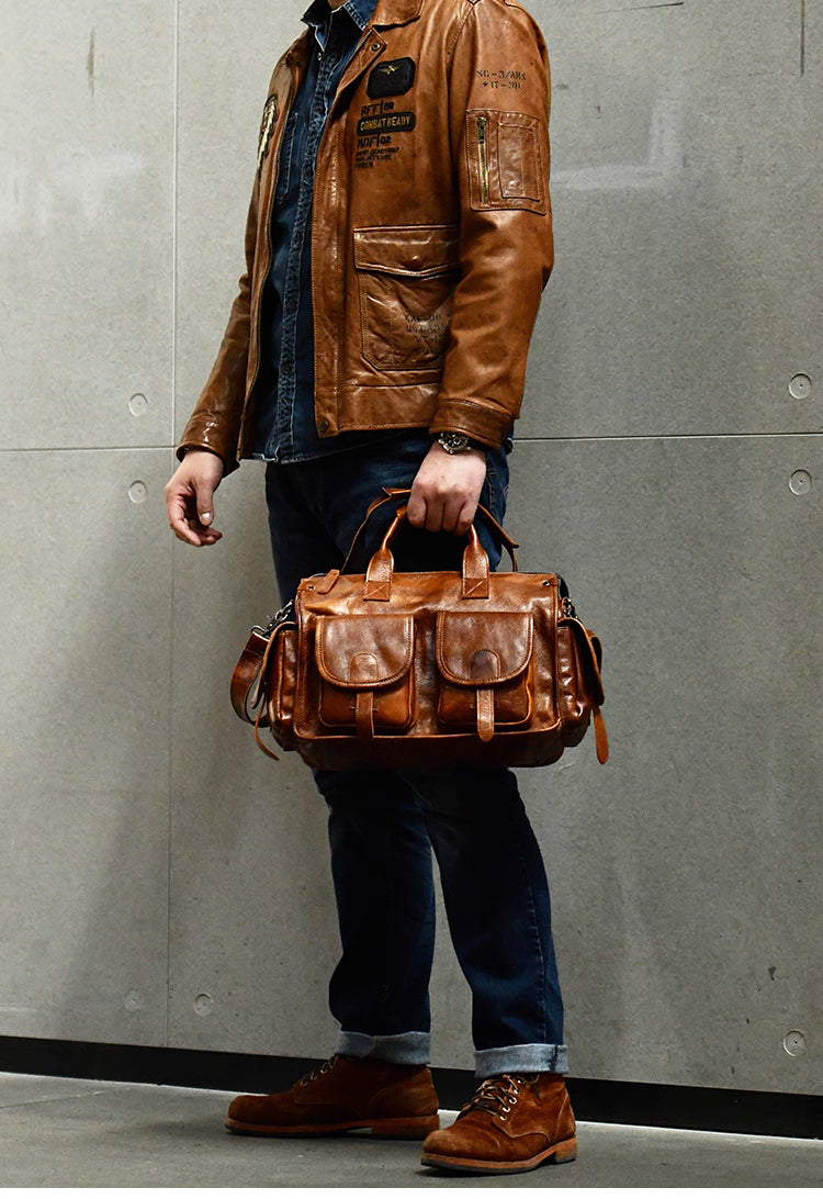 Man in denim and boots holding camel brown leather work bag in front of gray wall