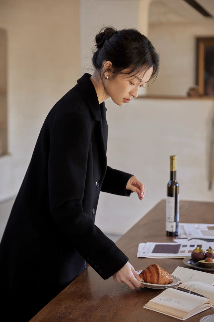Elegant woman in a black wool coat leaning over a table with a croissant and coffee in a cozy room.