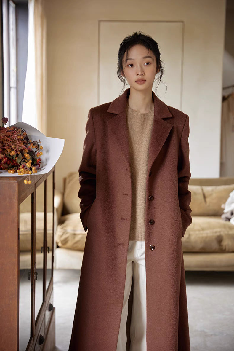 Woman in a warm brown wool long coat layered over a beige knit sweater and white pants, standing indoors.
