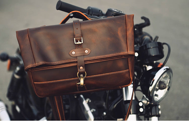 Close-up of the Nomad Crazy Horse Leather Messenger Bag hanging on a black motorcycle, showing rugged full-grain leather texture and brass hardware.