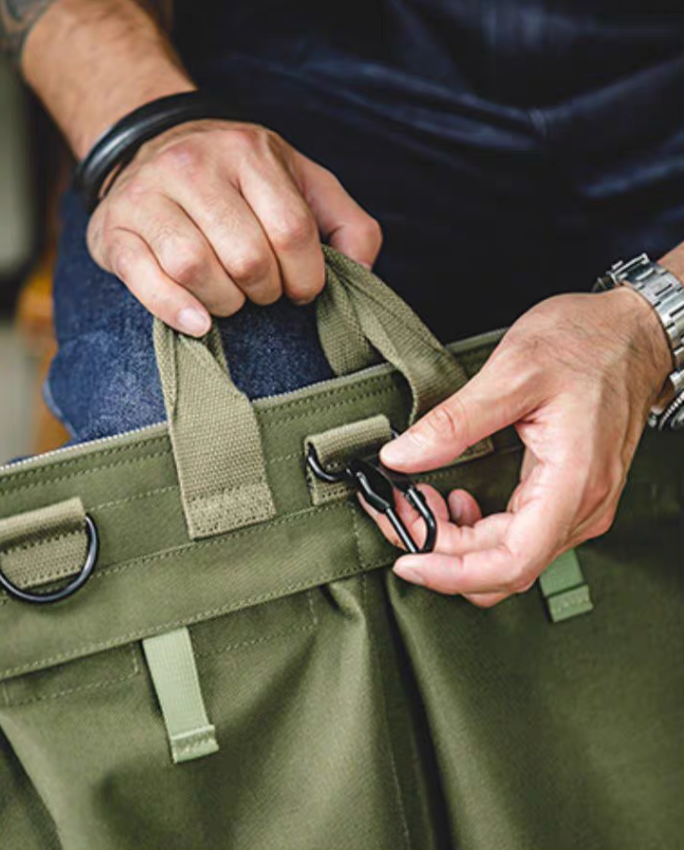 Close-up of army green canvas tote bag handle and front clip hardware detail with stitching and attachment points visible.