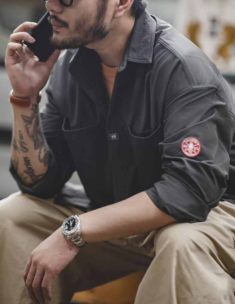 Lifestyle photo of man seated, wearing dark charcoal overshirt jacket with sleeve patch visible.