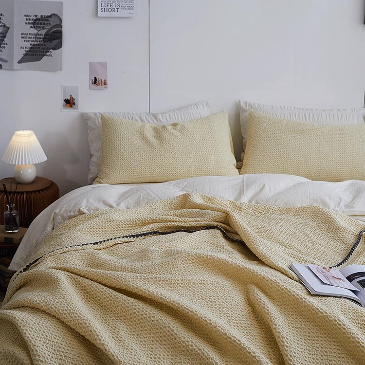 Light yellow 3D honeycomb waffle cotton throw blanket draped across a cozy bed with matching pillows in a minimalist bedroom setting.