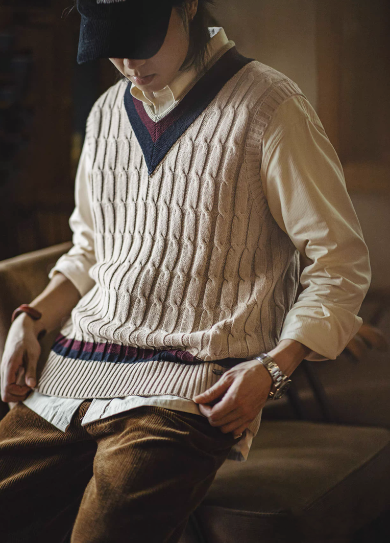 Man seated indoors wearing Ivy-style khaki V-neck cable knit vest layered over shirt