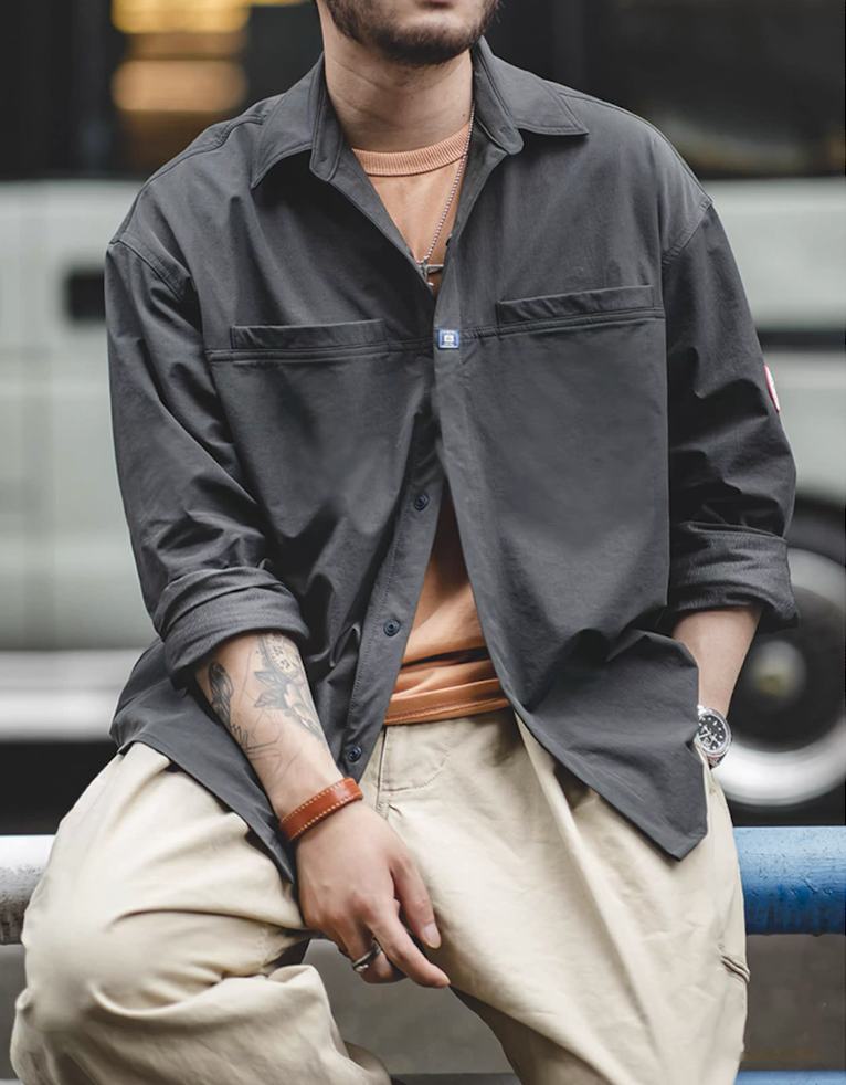 Model styling men’s dark charcoal overshirt jacket open with layered shirt and beige pants.