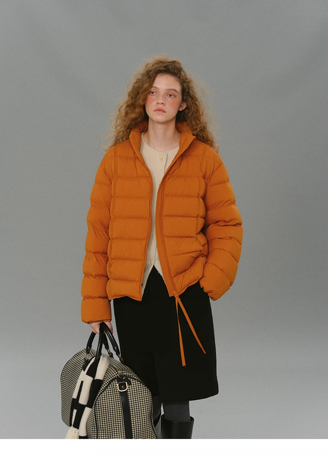 Model wearing a warm orange matte down puffer holding a checkered duffle bag.