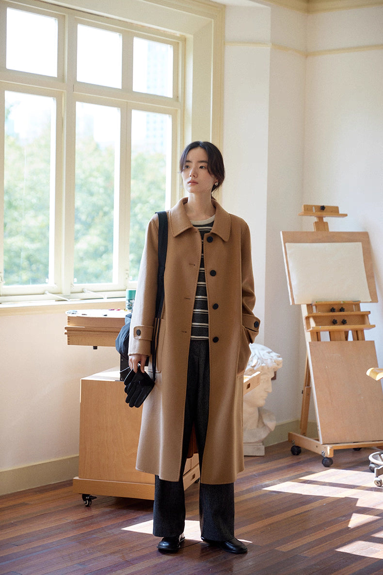 Full-length front view of almond-camel cashmere wool long coat, model standing by large window in art studio