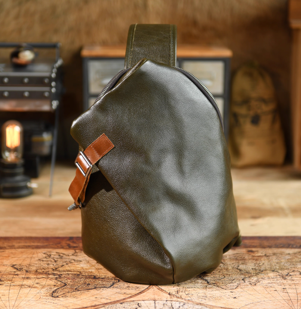Front view of army green full-grain leather sling bag with tan buckle strap on wooden table background.