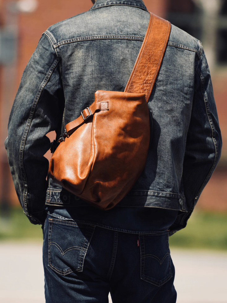 Man in denim jacket wearing caramel leather sling bag across the back outdoors.