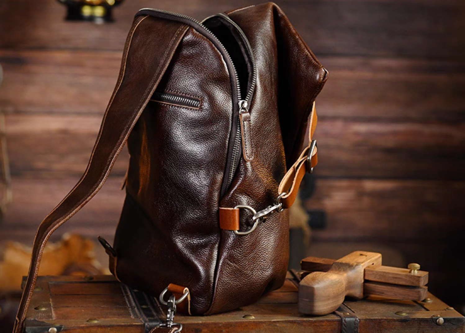 Side view of dark brown full-grain leather sling bag showing open zipper and tan strap buckle on rustic background.