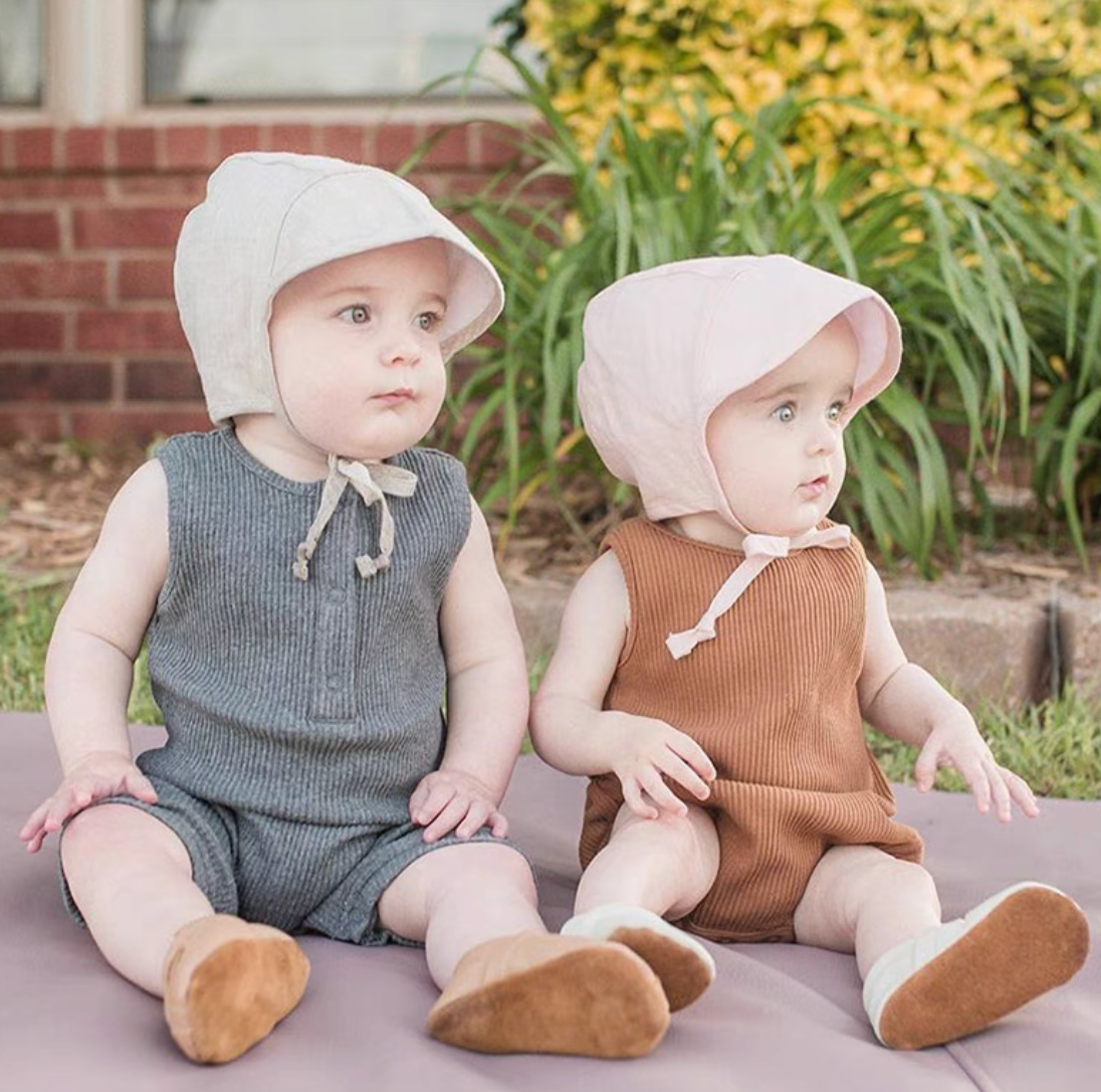 Two babies wearing linen cotton sun hats in cream and blush tones sitting outdoors together