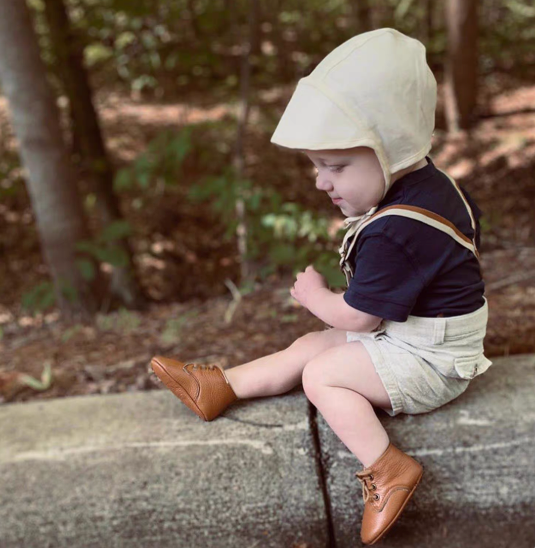 Baby wearing cream linen cotton sun hat sitting outdoors on stone ledge in forest setting
