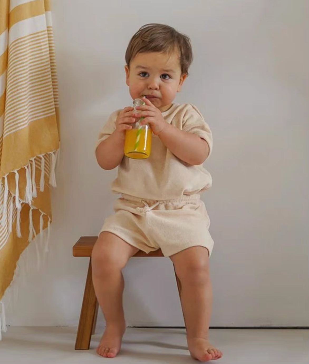 Baby in cream lounge set sitting on wooden chair drinking juice relaxed lifestyle scene