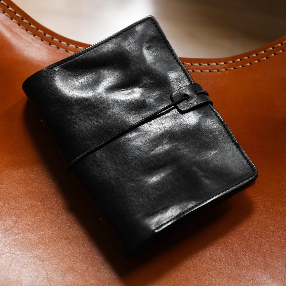 Classic black full-grain leather journal with elastic closure on tan leather chair.