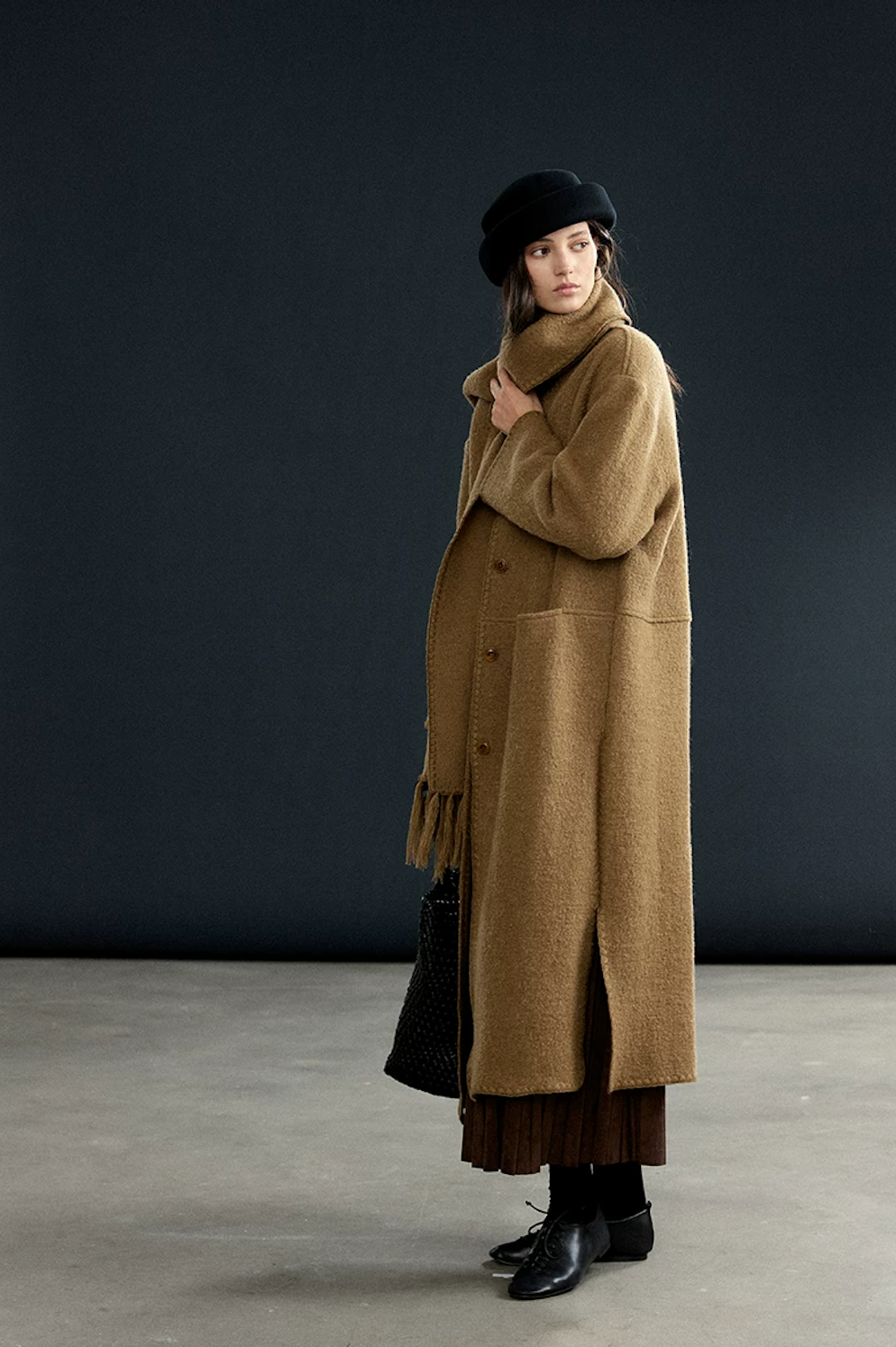 Camel long wool coat with matching fringed scarf wrapped high, showing blanket-stitch edging and side slit