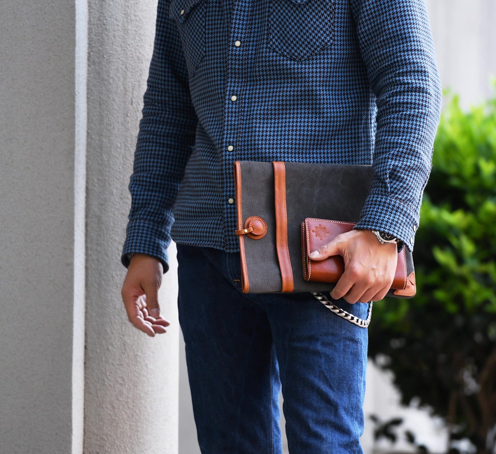 Man in blue patterned shirt carrying a charcoal gray canvas laptop sleeve with tan leather strap and corners.