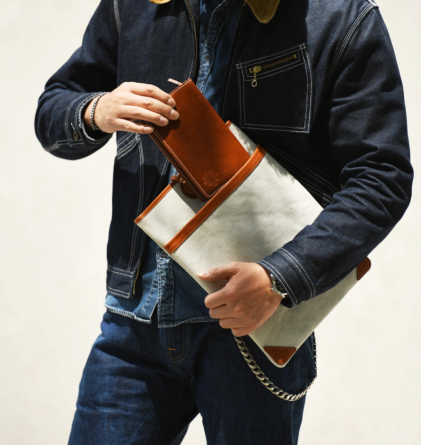 Man in dark denim jacket holding an ivory canvas laptop sleeve with brown leather trim and inserting a long wallet.