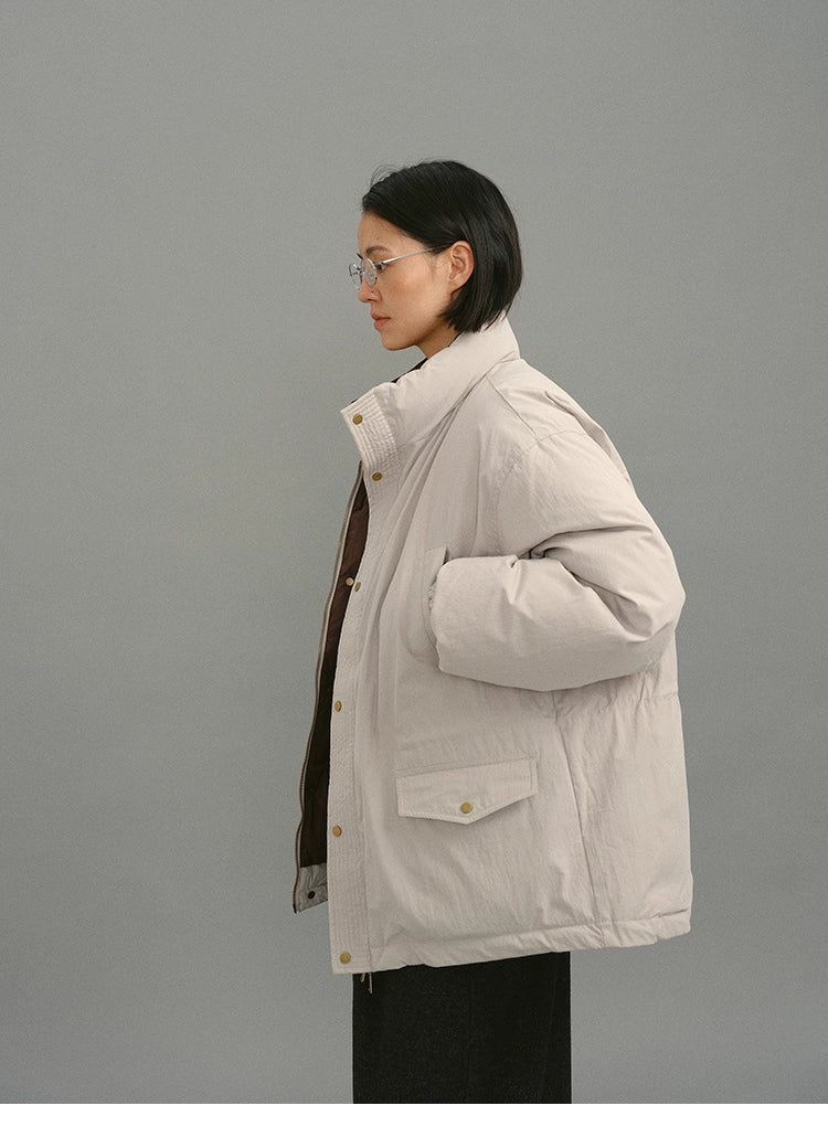 Side-profile of model wearing light greige down coat with hands in pockets and raised collar.