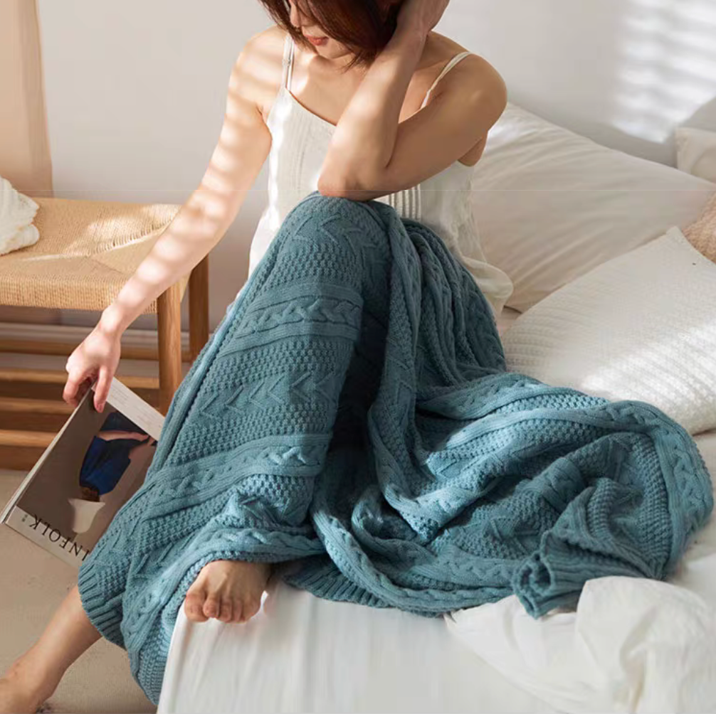 Green braided knit oversized throw blanket draped over bed with woman reading creating a relaxed cozy bedroom atmosphere