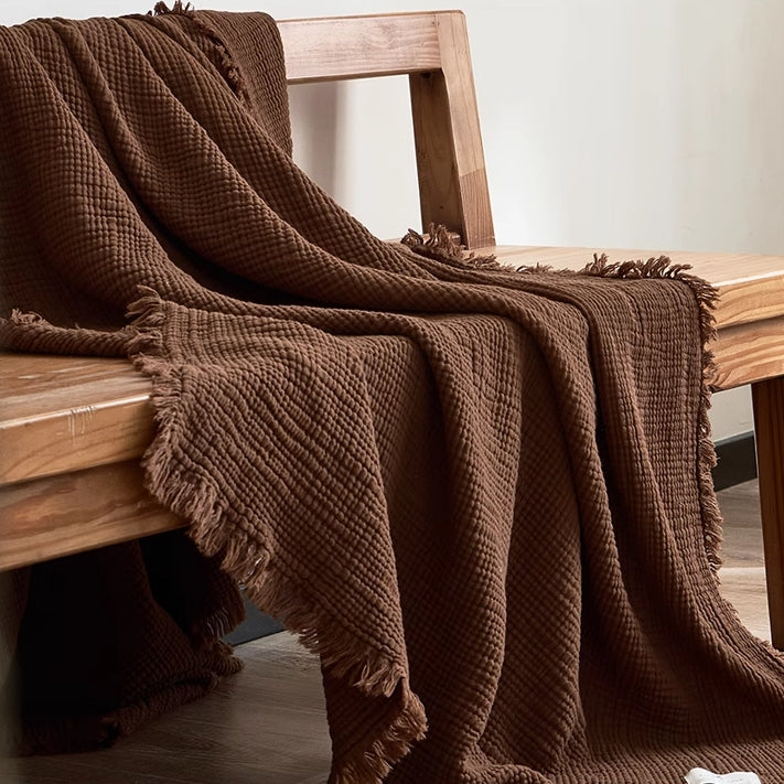 Chocolate brown waffle textured organic cotton fringe throw blanket draped over wooden chair in minimalist interior.