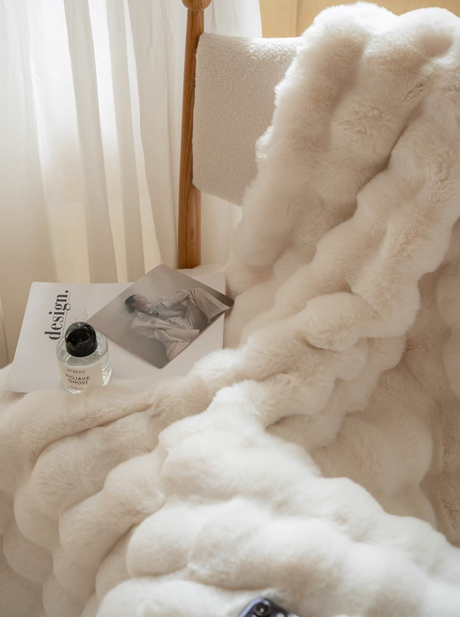 White plush faux fur throw blanket draped over chair with magazine and perfume, cozy modern interior styling