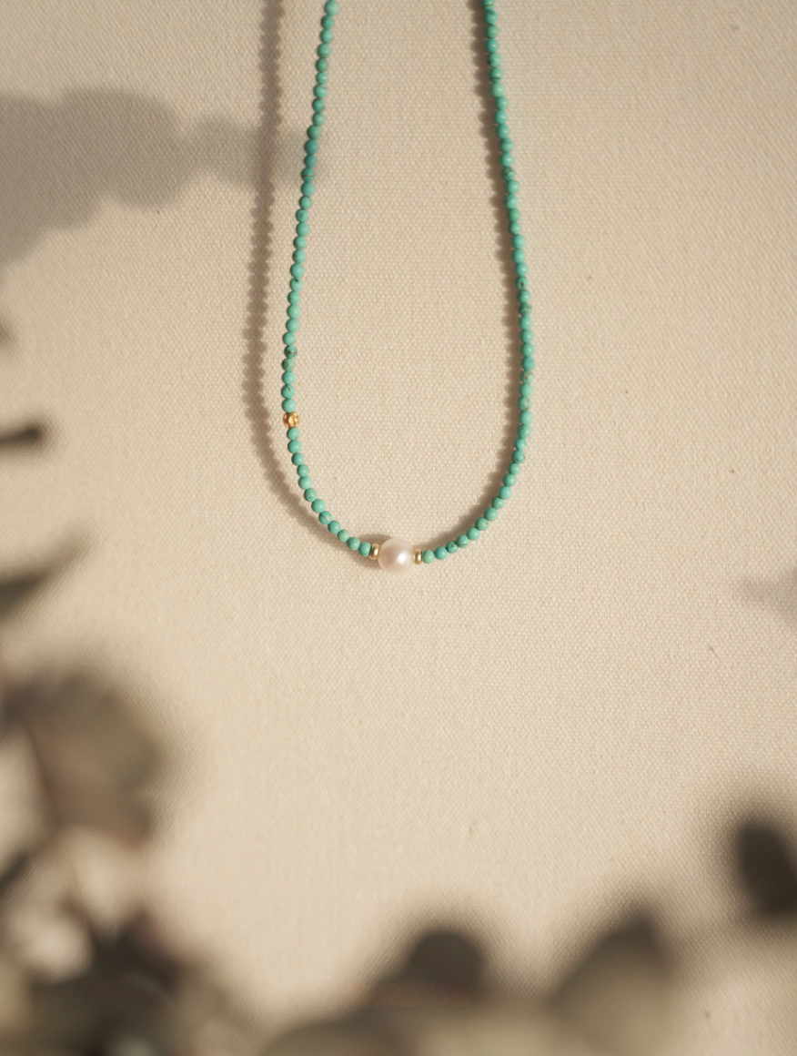Close-up of micro turquoise bead choker with freshwater pearl on textured fabric background with warm lighting