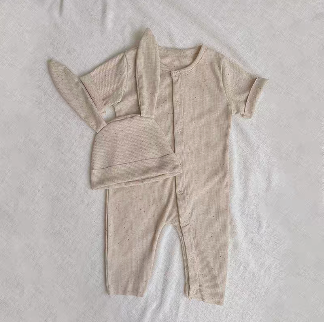 Oatmeal beige short sleeve long baby romper with bunny ear hat laid flat on fabric