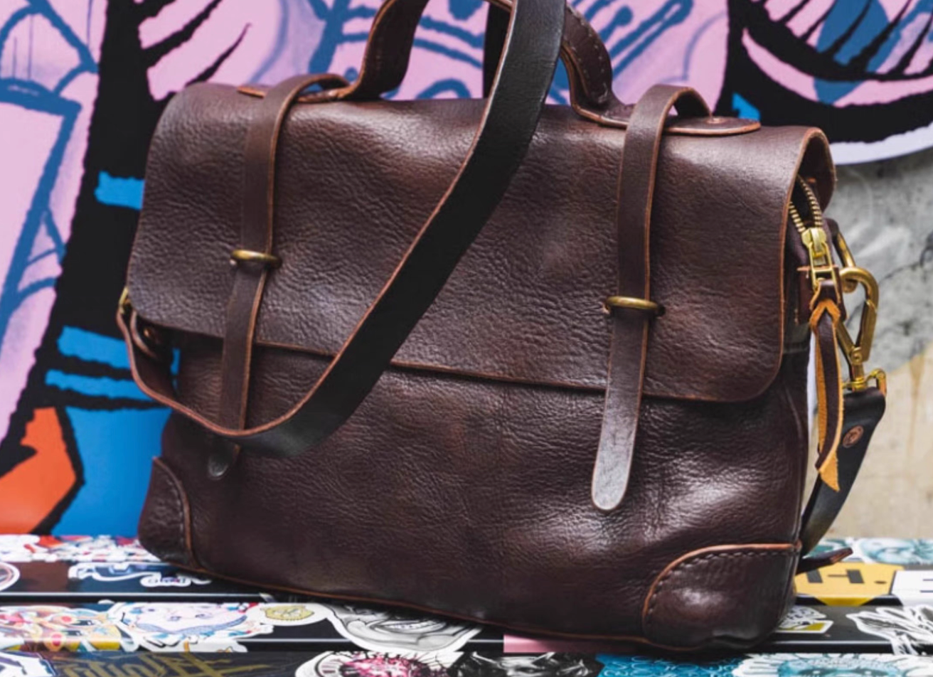 Vintage dark brown leather messenger bag with brass hardware placed against colorful graffiti wall