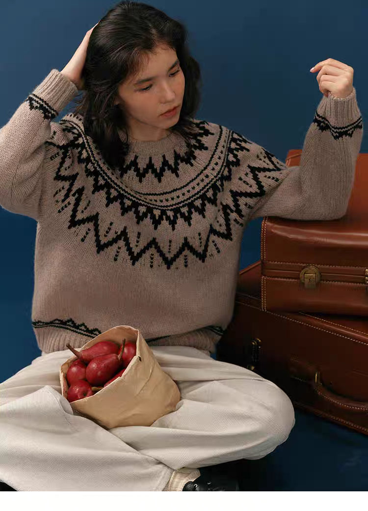 Woman sitting cross-legged in a taupe and black Fair Isle merino wool sweater with a paper bag of pears on her lap and vintage luggage beside her.