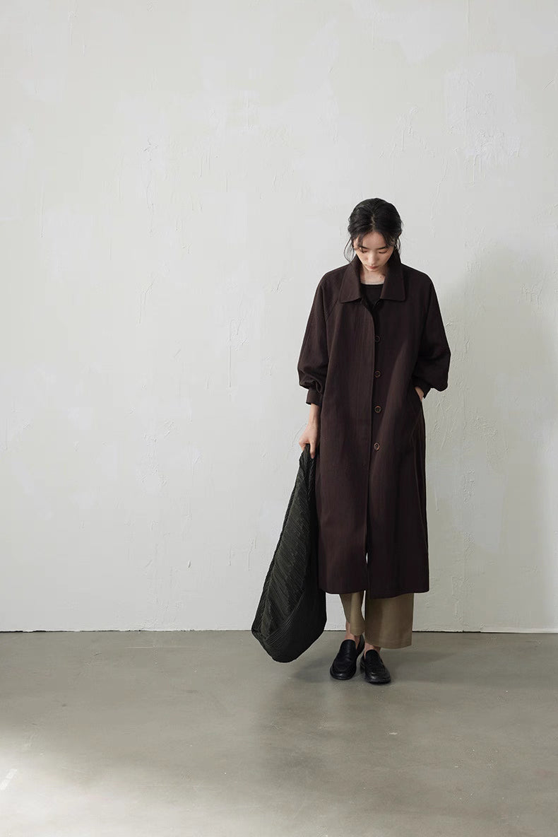Full-length centered front view of the coffee brown Tencel trench coat, model looking down with bag at her side.