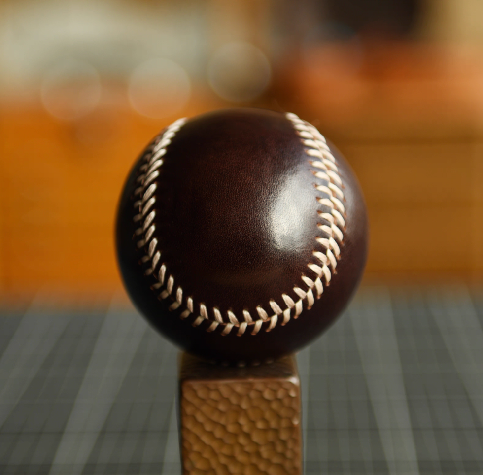Dark brown leather baseball on stand with visible glossy finish and white stitching.