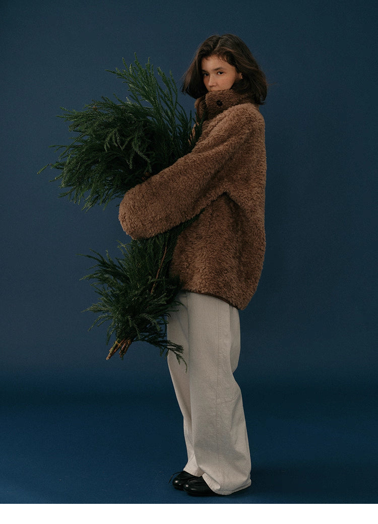 Full-body side view of model in chocolate teddy coat carrying large green winter branches.