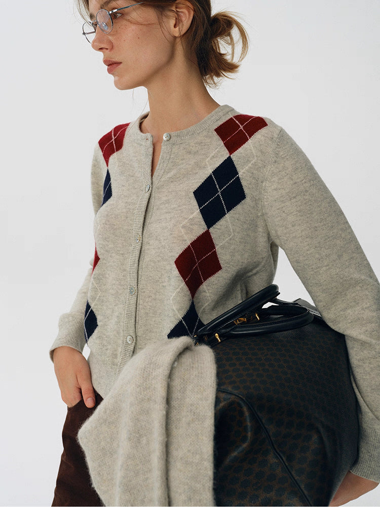 Side close-up of oat beige argyle wool cardigan, paired with a dark patterned travel bag and brown trousers.