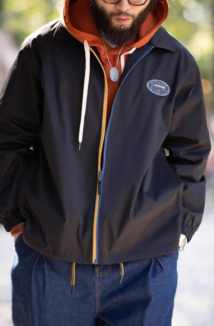 Man wearing black coach jacket over hoodie, styled with jeans outdoors.