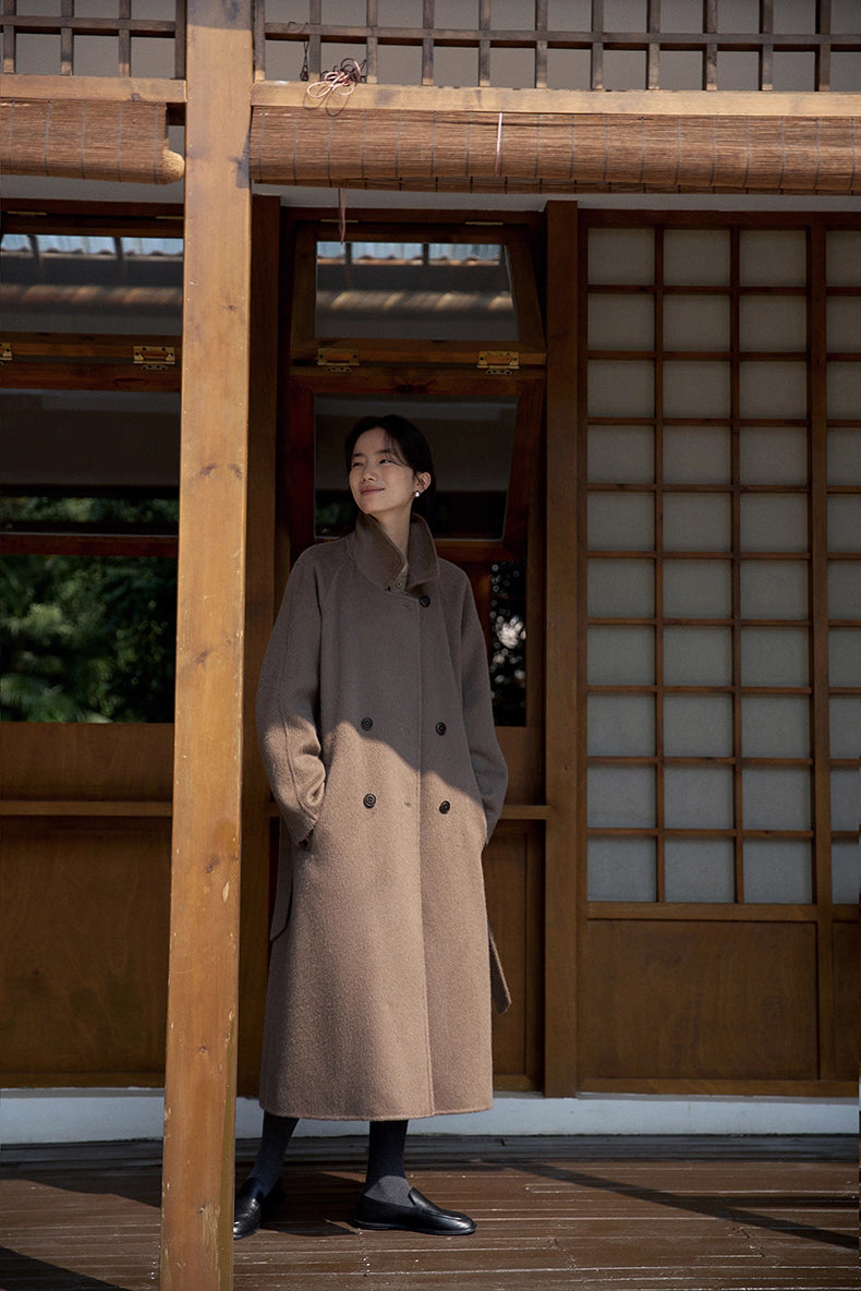 Full body image of woman standing on a wooden porch in a long taupe llama wool coat