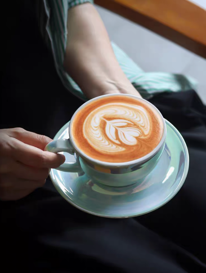 Luminous iridescent blue ceramic coffee cup and saucer with latte art held in hand creating a warm lifestyle coffee moment