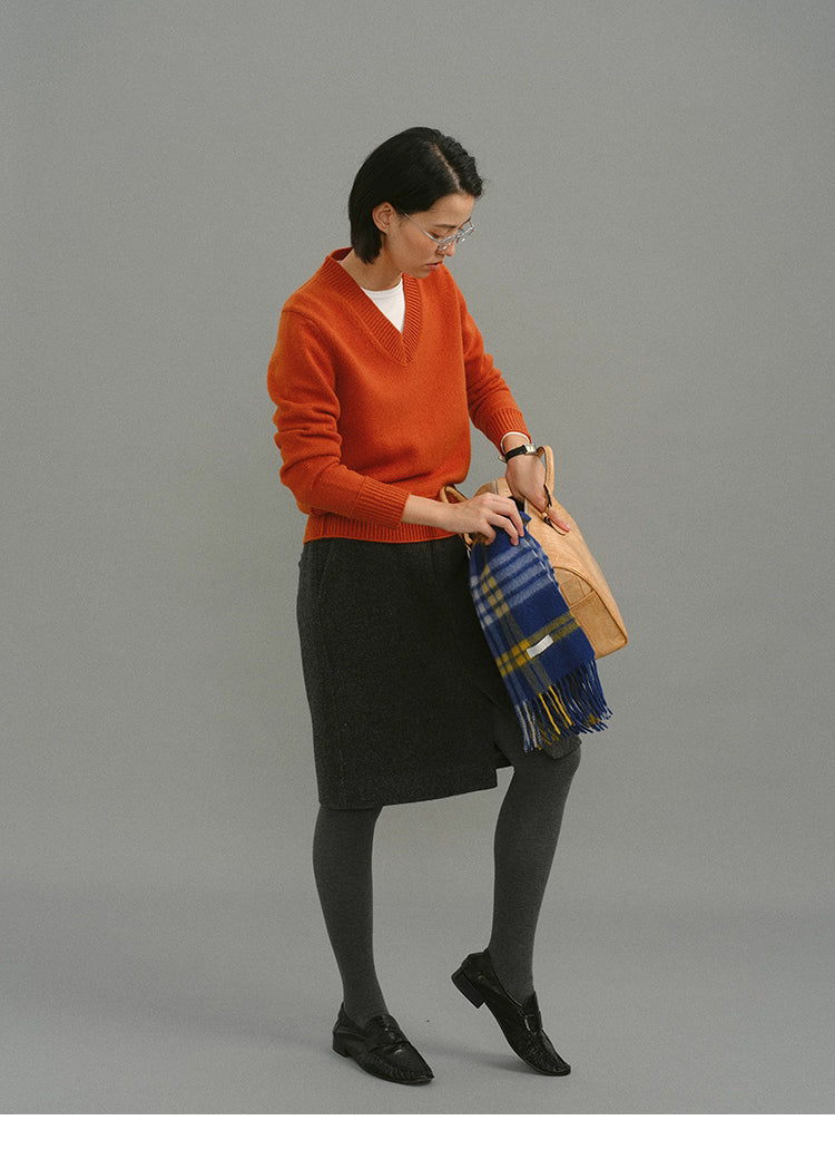 Model adjusting a tan handbag with a blue plaid scarf while wearing a rust-orange Merino V-neck sweater and charcoal midi skirt.