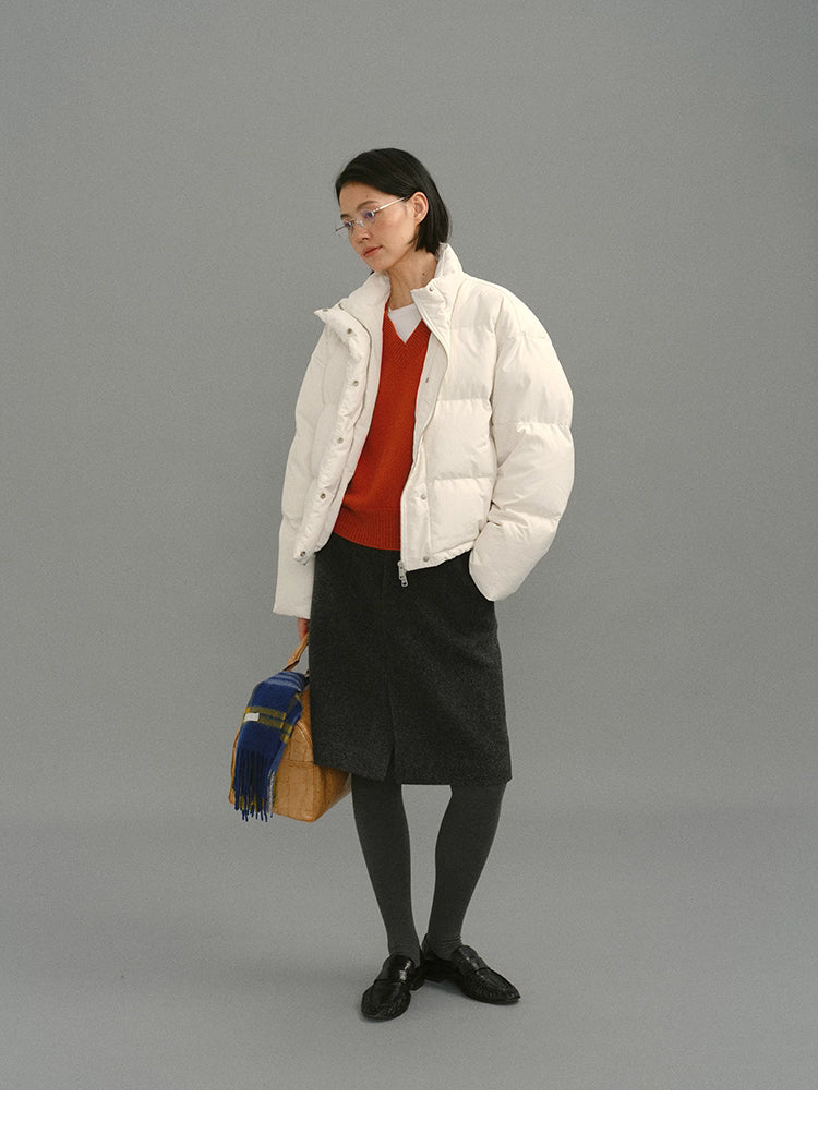 Full-body shot of model wearing a white puffer jacket layered over a rust-orange V-neck Merino sweater, styled with a charcoal skirt, grey tights, and tan handbag with a plaid scarf.