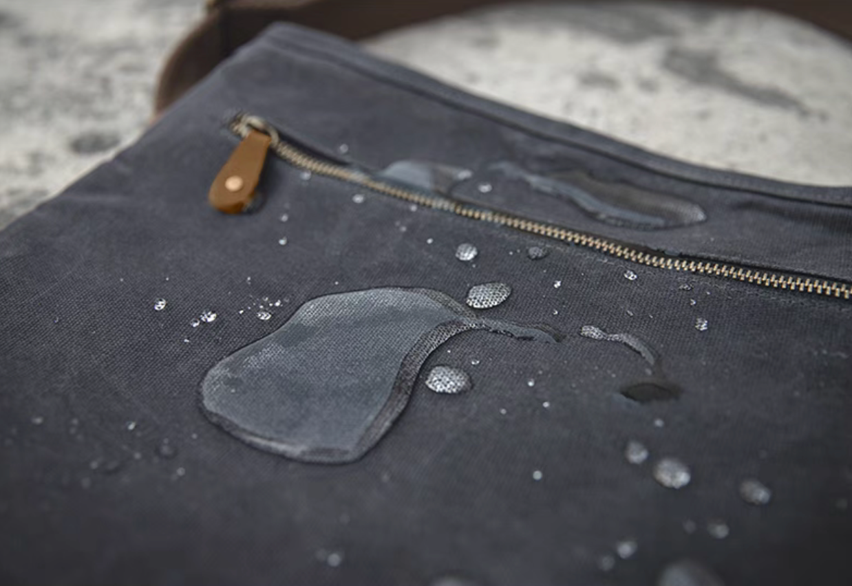 Dark grey waxed canvas crossbody bag surface with water droplets demonstrating water-resistant material and zipper detail.