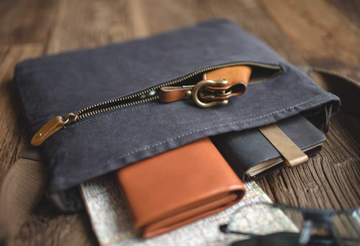 Dark grey waxed canvas crossbody bag partially unzipped showing interior storage with a wallet and notebook inside on a wooden background.