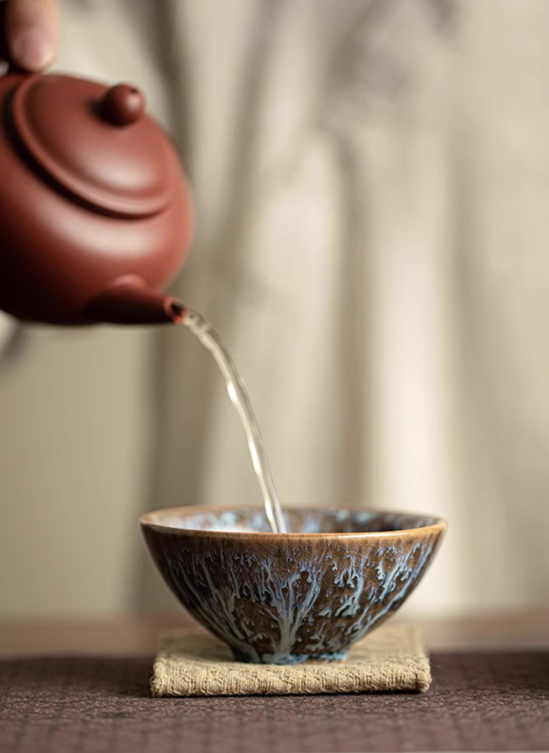 Close-up of tea being poured into dark kiln-glazed ceramic tea cup highlighting textured glaze surface