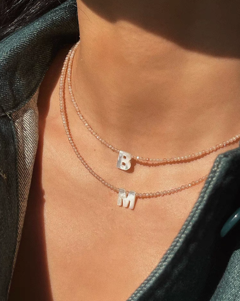 Layered personalized shell initial necklaces with letters B and M worn on neck in natural bead chain design