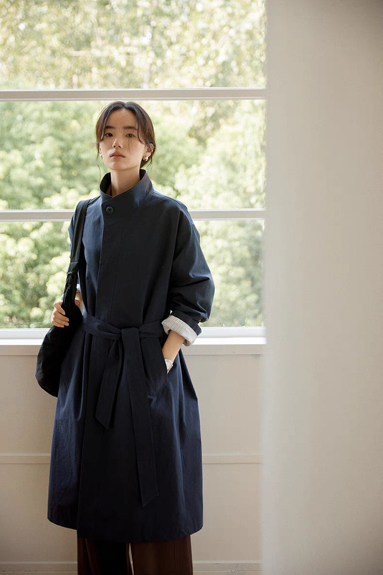 Woman wearing the blue Relaxed Cotton-Linen Trench in a three-quarter front view by the window, showcasing the belted waist, relaxed fit, and understated modern style