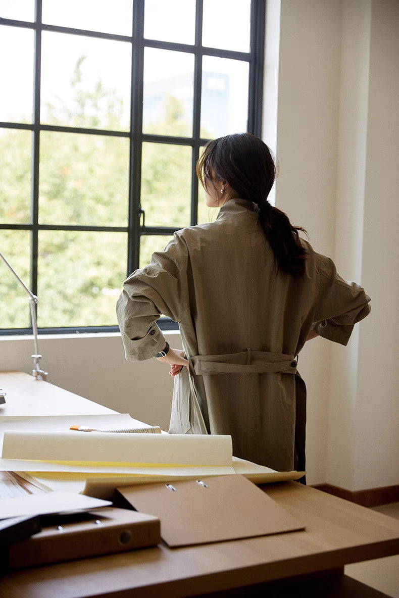 Back view of the gray Relaxed Cotton-Linen Trench showing the detachable belt, relaxed long silhouette, and minimalist tailoring near a sunlit window